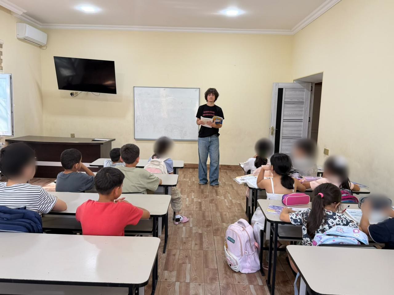 Jamal stands at the front of a classroom, reading aloud from a book to a group of children seated at desks.
