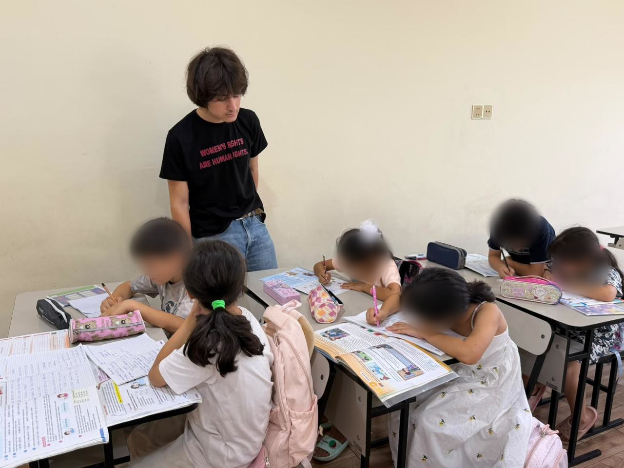 Jamal stands beside desks where children are writing in their exercise books.