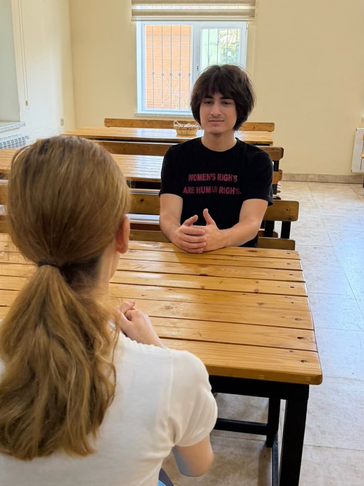 Jamal sits at a wooden table, listening to a woman across from him, who is seen from behind.
