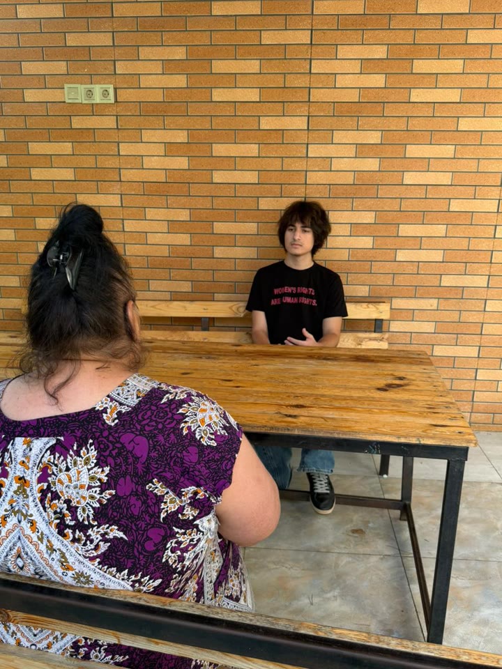 Jamal sits across a wooden table from a woman, who is seen from behind.